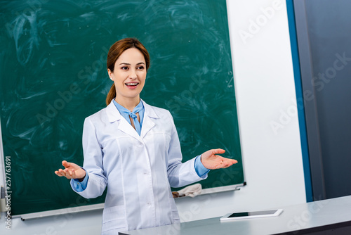 Smiling chemistry teacher in white coat explaining lesson in front of blackboard