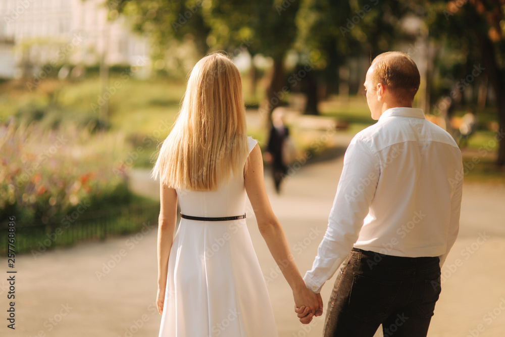 Young beautiful couple, husband in a white shirt and a wife in a dress walking around the park in summer time Young beautiful couple, husband in a white shirt and a wife in a dress walking around the park in summer time