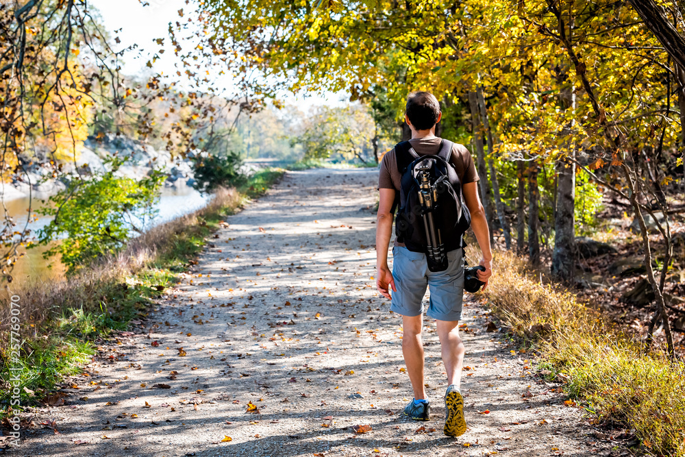 Obraz premium Young photographer man back walking on trail path road during autumn Potomac river in Great Falls, Maryland with colorful foliage and backpack tripod