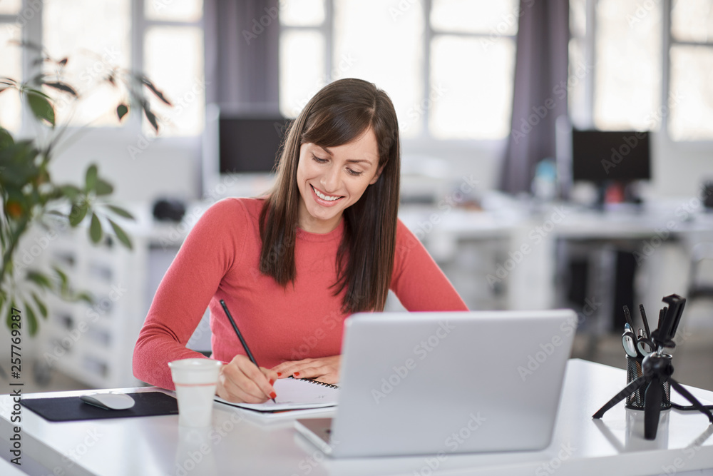 Charming Caucasian businesswoman sitting in moderna office and taking notes in notebook. In front of her laptop. Charming Caucasian businesswoman sitting in moderna office and taking notes in notebook. In front of her laptop.