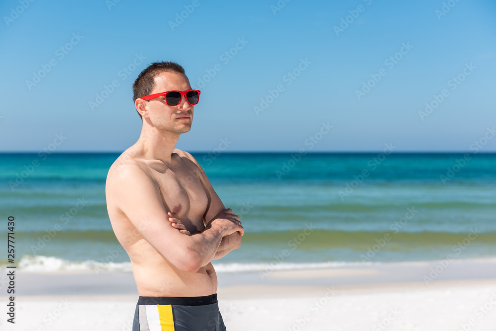 Young handsome attractive fit muscular man hipster millennial on beach during sunny day with red sunglasses in Florida panhandle ocean looking to side Young handsome attractive fit muscular man hipster millennial on beach during sunny day with red sunglasses in Florida panhandle ocean looking to side