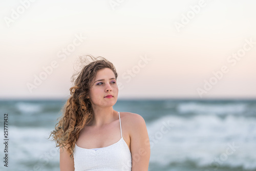 Young woman in white dress on beach sunset in Florida panhandle with hair blowing in wind and bokeh of ocean waves background looking up © Kristina Blokhin