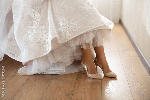Bride putting on wedding shoes at home where she's getting ready - Wearing white dress in a bright room with wooden floor