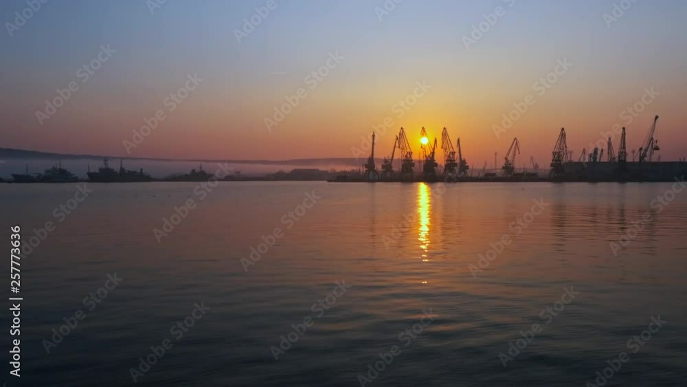 Maritime city port. Quiet sea surface disturbance. A flock of seabirds, gulls, circling over ships and cranes against the backdrop of a bright orange sunset Maritime city port. Quiet sea surface disturbance. A flock of seabirds, gulls, circling over ships and cranes against the backdrop of a bright orange sunset