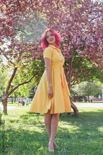 Beautiful and young woman with bright red hair and ref lips standing next to a blooming apple tree in a yellow dress. photo