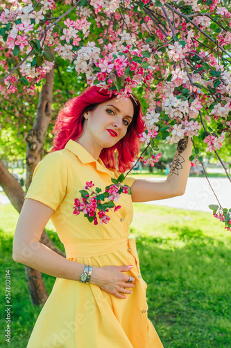 Beautiful and young woman with bright red hair and ref lips standing next to a blooming apple tree in a yellow dress.