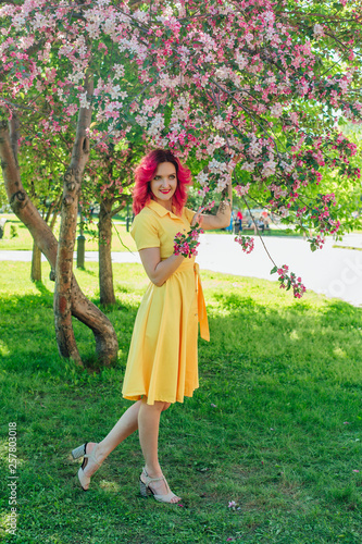 Beautiful and young woman with bright red hair and ref lips standing next to a blooming apple tree in a yellow dress. photo