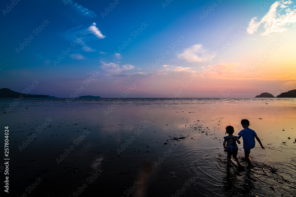 Far-away view of a muddy shore at low tide with children with blue and orange gradient sky and wispy clouds