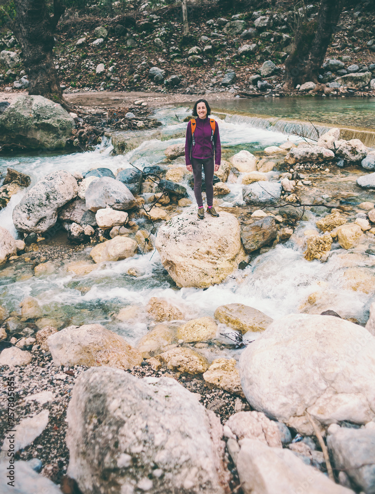 A woman with a backpack is standing near a mountain river.