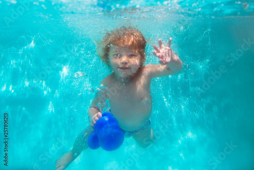 Smiling baby girl in cute modern dress diving underwater in blue swimming pool. Active lifestyle, child swimming lesson with parents. Water sports activity during family summer vacation in resort