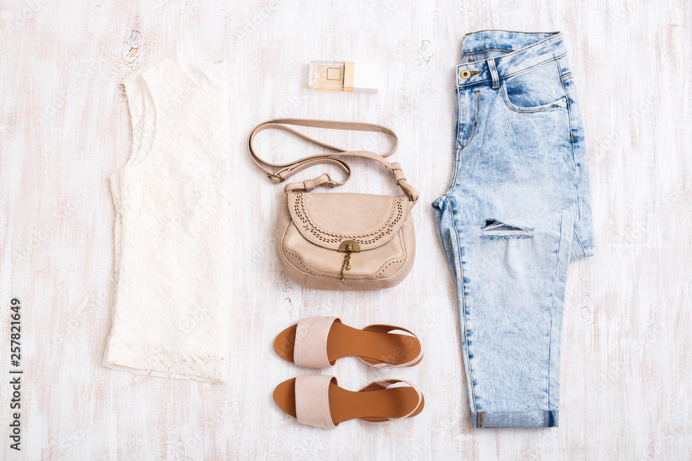 White lace top, light blue ripped boyfriend jeans, beige sandals, perfume,  small beige leather bag on white wooden background. Overhead view of  woman's casual outfits. Flat lay, top view. Stock Photo