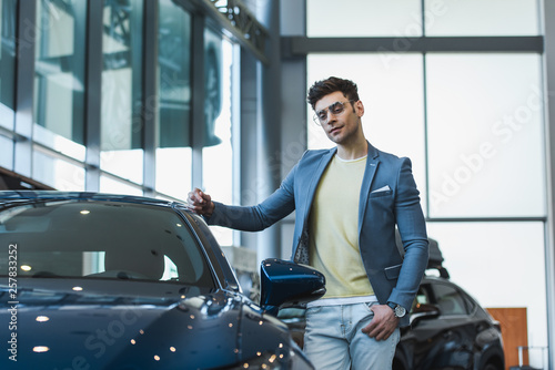 pensive man in glasses standing with hand in pocket near automobile in car showroom
