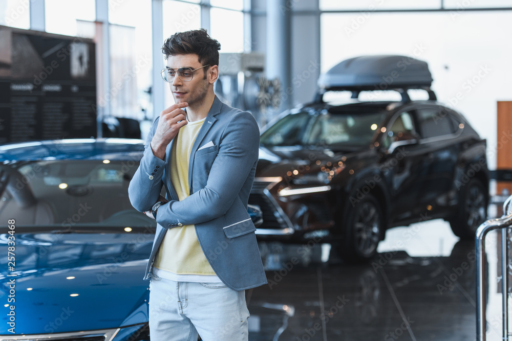 thoughtful man in glasses standing near automobiles in car showroom thoughtful man in glasses standing near automobiles in car showroom