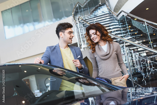 stylish man in glasses pointing with finger at car near curly attractive woman standing with hand in pocket