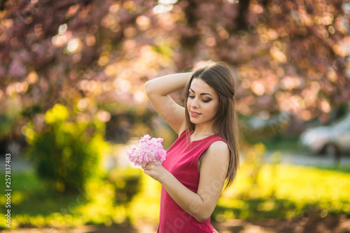 Fototapeta Naklejka Na Ścianę i Meble -  Charming girl in beautiful pink dress hold bunch of sakura in hands. Beautiful european girl near the japanese tree