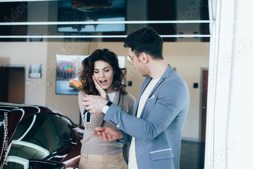 cheerful man holding keys near surprised curly girl while standing near red automobile