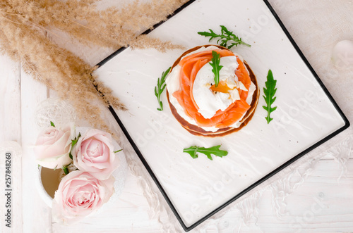 Beautiful pancakes with salmon, poached egg and arugula on a white plate on a light background, decorated with pink roses. Close-up. Space. Top view. Flat lay