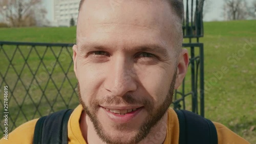 portrait of a bearded young man who pensive looking into camera and then smiling and looks away on the sun on summer city background slow motion close up video in 4K