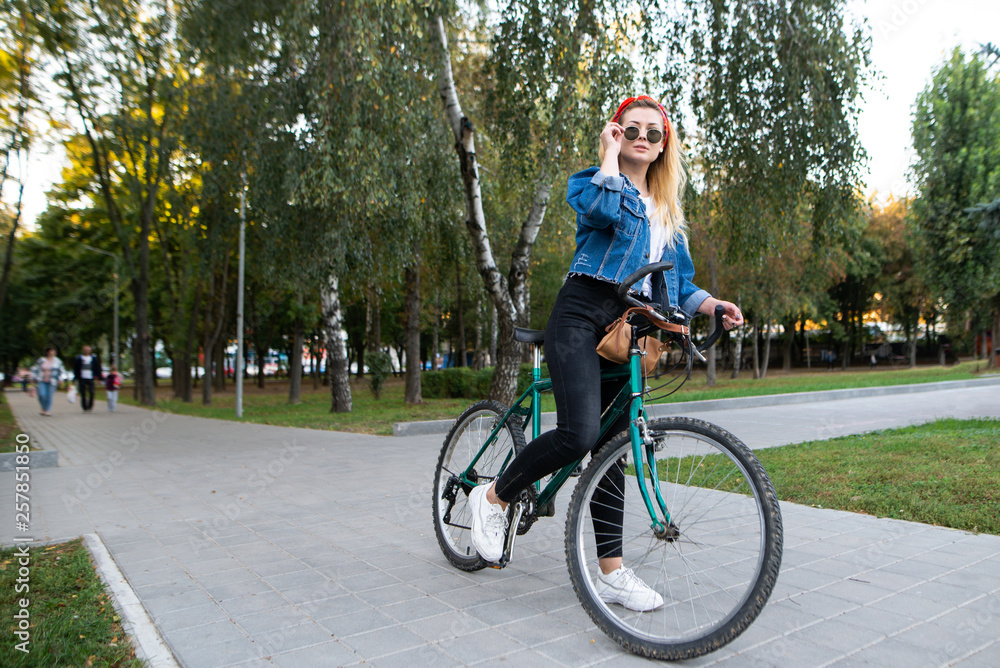 Stylish attractive woman wearing sunglasses sits on a bike in the park. Hipster girl rides a bike in the park. Active rest