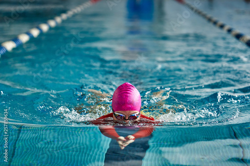 Female athlete in a red-yellow swimsuit is swimming in the style of breaststroke. Splashes of water scatter in different directions.