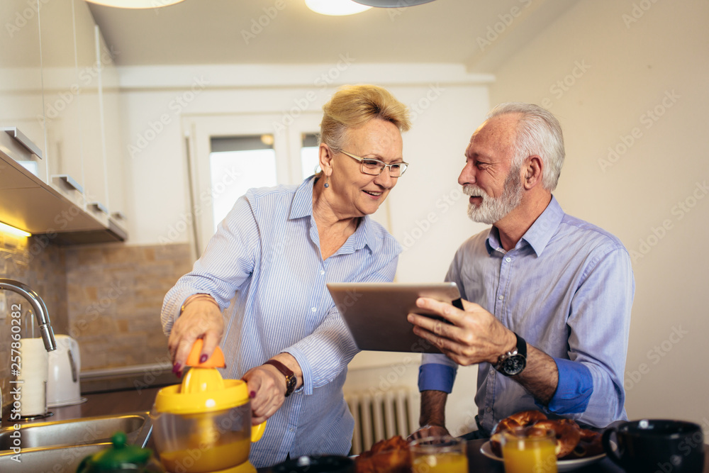 Aged couple busy look at digital tablet while having delicious breakfast at home kitchen Aged couple busy look at digital tablet while having delicious breakfast at home kitchen