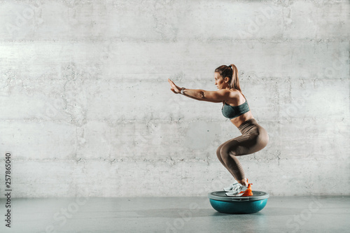 Side view of muscular Caucasian woman in sportswear and with ponytail doing squat endurance on bosu ball. In background wall. photo