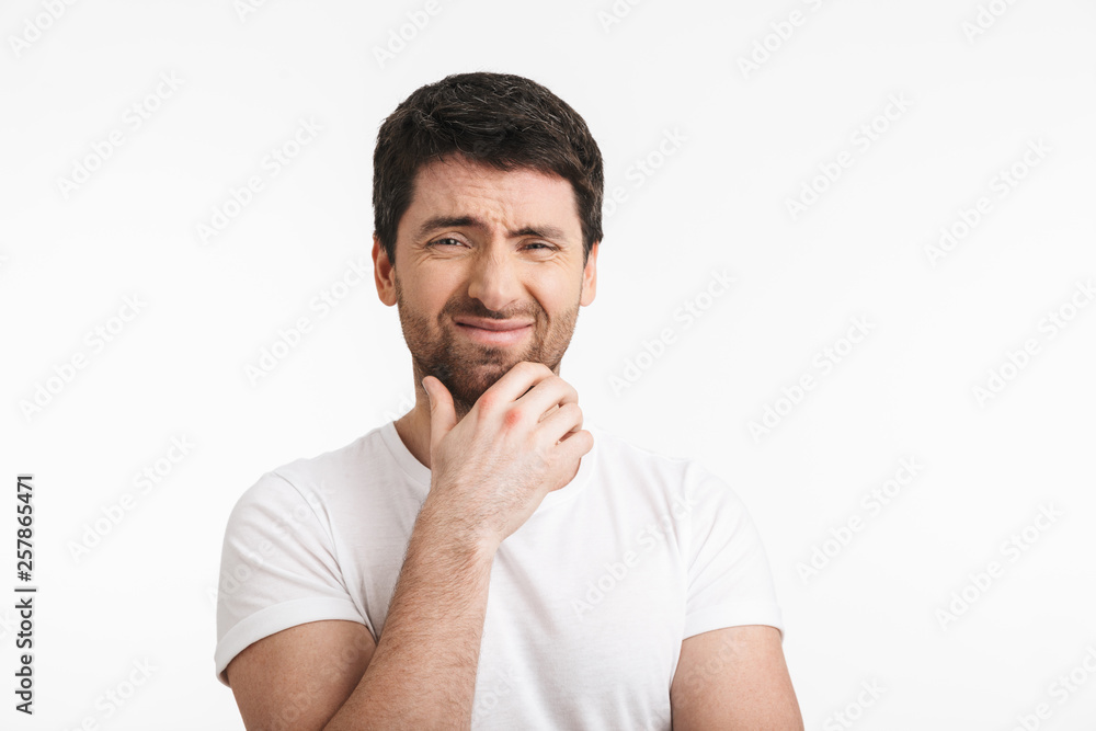 Image of frowning man 30s with bristle in casual t-shirt thinking and touching chin Image of frowning man 30s with bristle in casual t-shirt thinking and touching chin