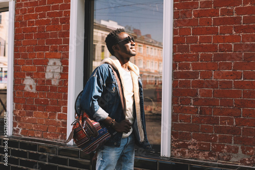 Wallpaper Mural Happy smiling african man in jeans jacket, with backpack looking up at sunlight walking on city street over brick wall background Torontodigital.ca