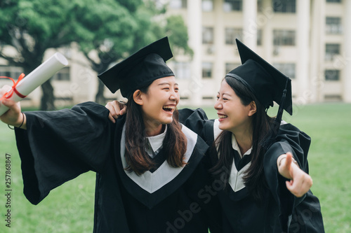 Graduation asian female friends hold diploma. cheerful ladies students hugging shoulders arms with thumb up finger hand gesture best sign. beautiful cheerful women students looking each other smiling photo