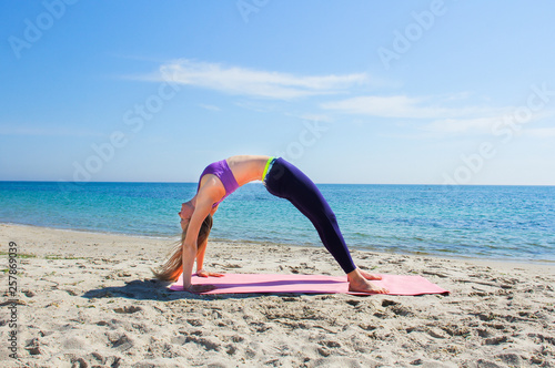 Woman training yoga on the beach. Girl is wearing sportive clothes doing sport on mat on the sea beach. Caucasian female practicing fitness, pilates and meditation. 