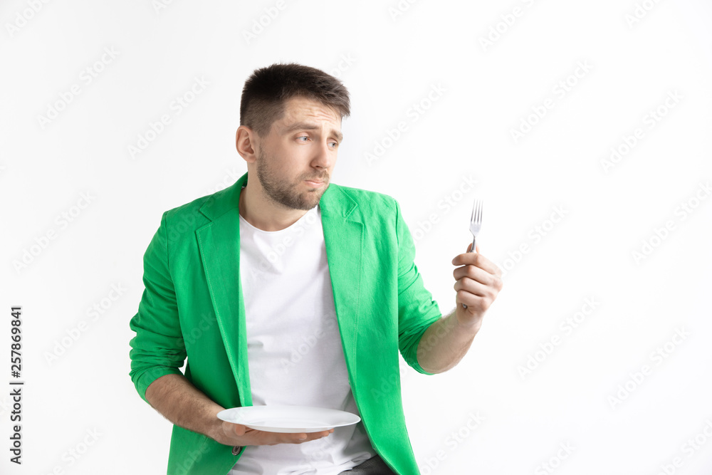 Young sad attractive caucasian guy holding empty dish and fork isolated on grey background. Copy space and mock up. Blank template background. Reject, rejection concept Young sad attractive caucasian guy holding empty dish and fork isolated on grey background. Copy space and mock up. Blank template background. Reject, rejection concept
