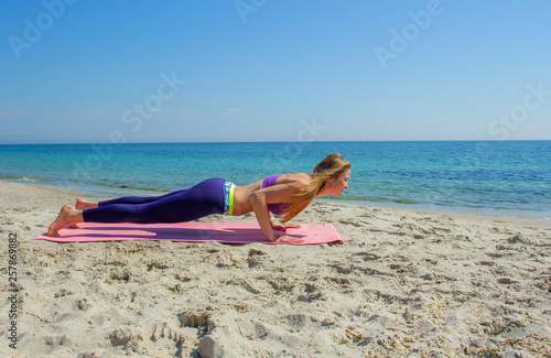 Woman training yoga on the beach. Girl is wearing sportive clothes doing sport on mat on the sea beach. Caucasian female practicing fitness, pilates and meditation. 