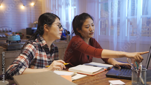 two serious asian woman discussing on project report for final exam. girl pointing laptop screen showing classmate something talking. group of college students teamwork on kitchen table in dark home.