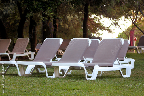 Exotic beach on mediterranean sea, sunbeds for sunbathing and relax on grass in tropical garden of luxury resort hotel. Sun loungers on lawn waiting for tourists. Idyllic seaside in summer season.