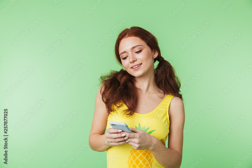 Beautiful young redhead girl posing isolated over green wall background using mobile phone. Beautiful young redhead girl posing isolated over green wall background using mobile phone.
