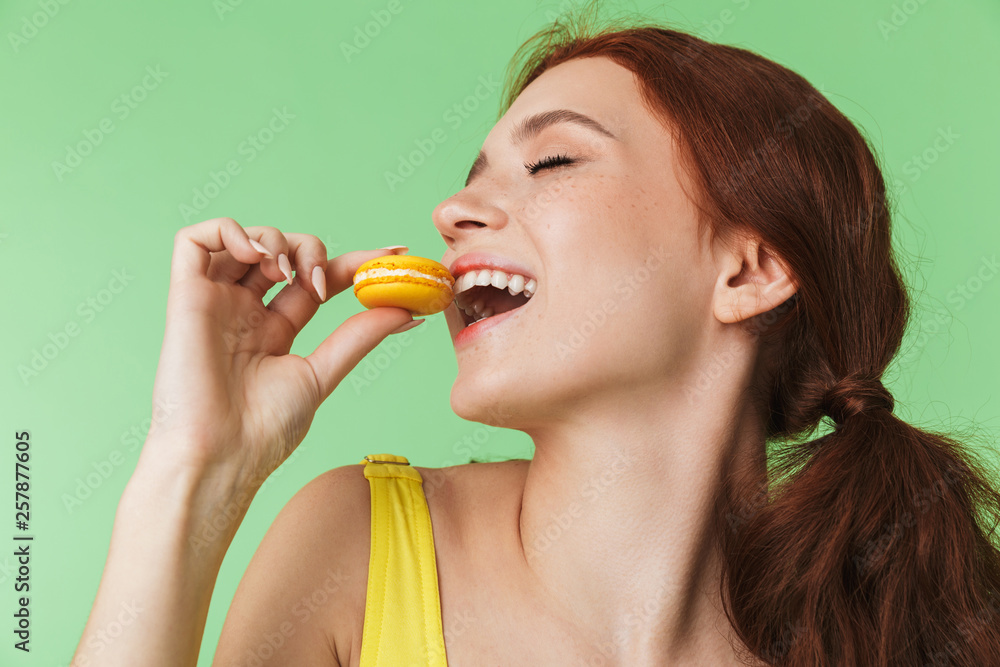 Redhead girl posing isolated over green wall background holding sweeties macaroons. Redhead girl posing isolated over green wall background holding sweeties macaroons.