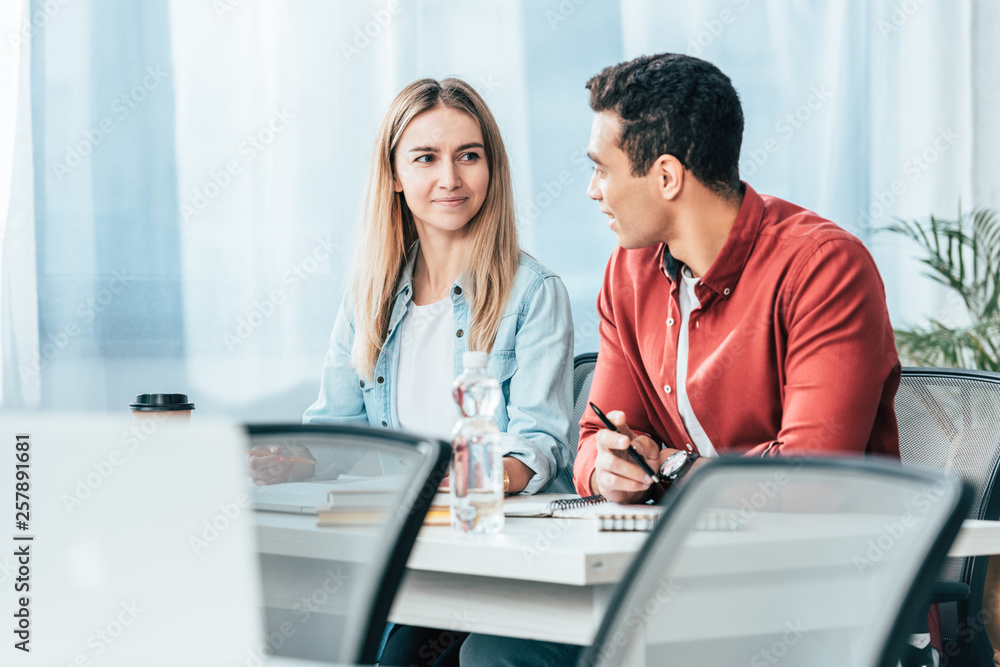 Multicultural students in casual clothes sitting at desk and looking at each other Multicultural students in casual clothes sitting at desk and looking at each other