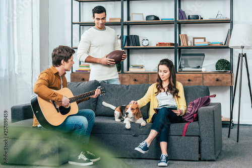 Three multicultural friends with beagle dog playing guitar in living room