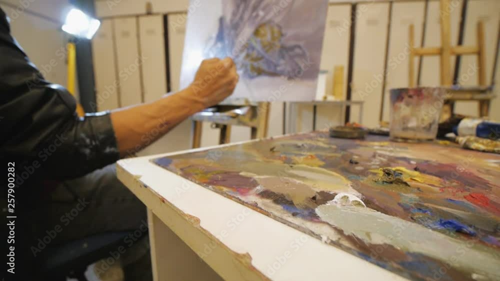 Male hispanic artist in black leather jacket paints oil still-life on canvas sitting on chair in art studio. Focused on painting colors. Male hispanic artist in black leather jacket paints oil still-life on canvas sitting on chair in art studio. Focused on painting colors.