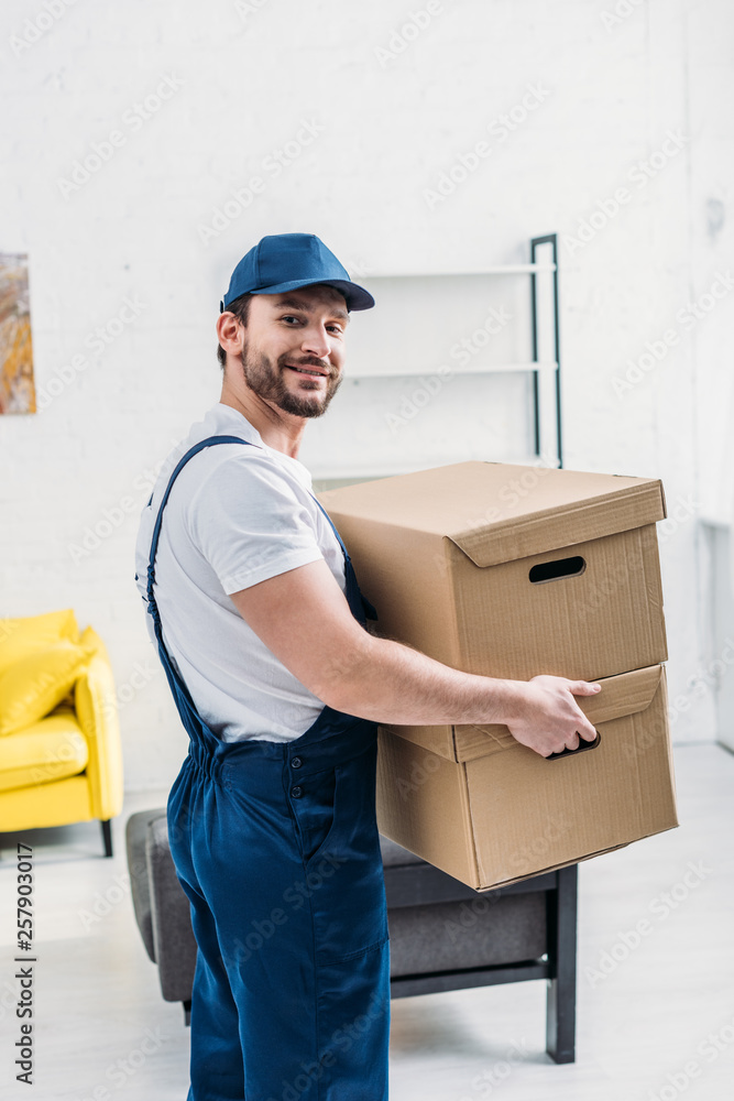 handsome mover in uniform looking at camera while carrying cardboard boxes in apartment handsome mover in uniform looking at camera while carrying cardboard boxes in apartment
