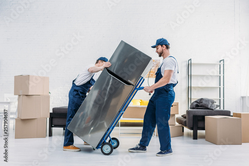 Fototapeta two movers in uniform using hand truck while transporting refrigerator in apartm