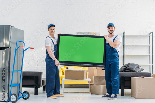 two movers in uniform looking at camera while transporting tv with green screen in apartment