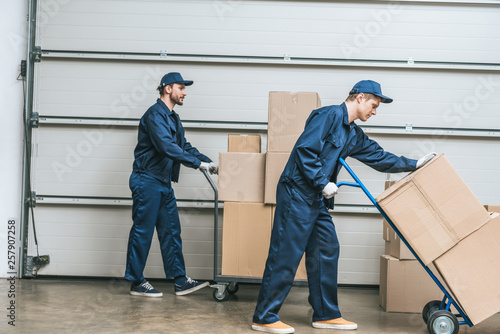 Canvas Print two movers in uniform using hand trucks while transporting cardboard boxes in wa