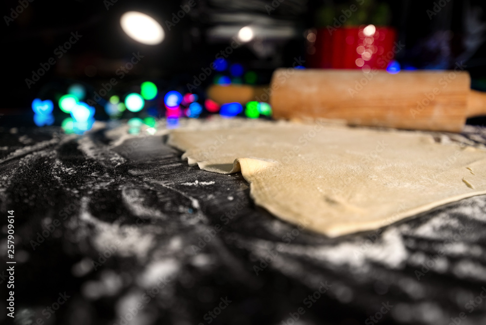 Uncooked rolled dough on table in kitchen. Christmas or fairy lights and decorations in background. Flour all around. Baking gingerbread, cookies, bread or pastry Uncooked rolled dough on table in kitchen. Christmas or fairy lights and decorations in background. Flour all around. Baking gingerbread, cookies, bread or pastry