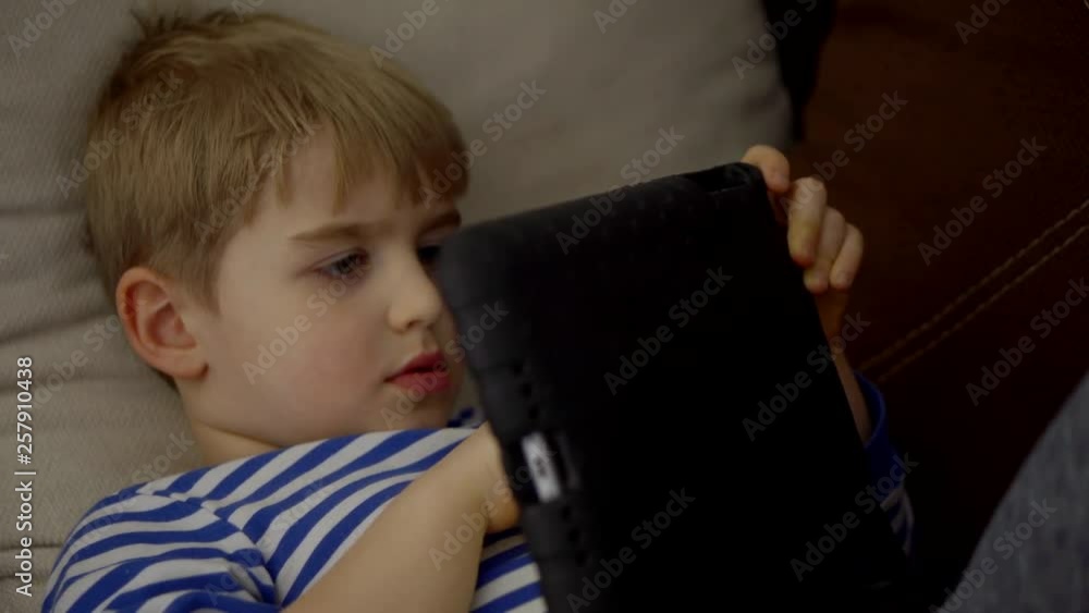 European baby boy sitting in bed, playing with tablet PC. Children playing with tablet computer, gadget-dependent children, the concept of pseudo-autism European baby boy sitting in bed, playing with tablet PC. Children playing with tablet computer, gadget-dependent children, the concept of pseudo-autism