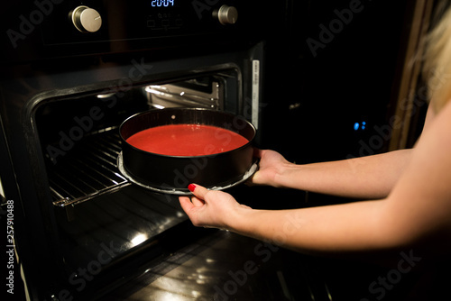 Woman is cooking in kitchen, baking a red velvet cake. Taking out or putting a biscuit tray filled with dough in oven. Delicious, homemade dessert