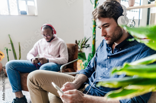 Two young men with cell phones and headphones sitting in armchairs in a loft