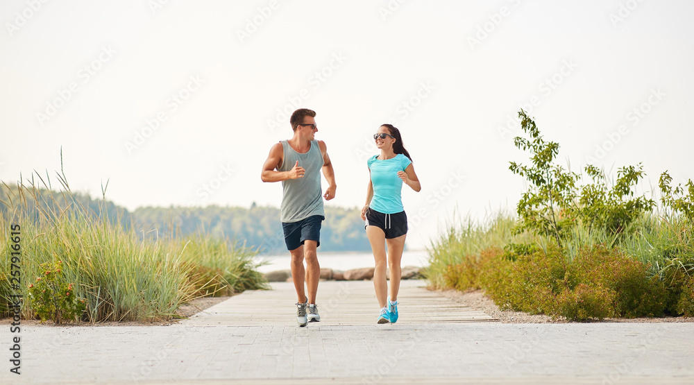 fitness, sport and lifestyle concept - happy couple in sports clothes and sunglasses running along summer beach path fitness, sport and lifestyle concept - happy couple in sports clothes and sunglasses running along summer beach path