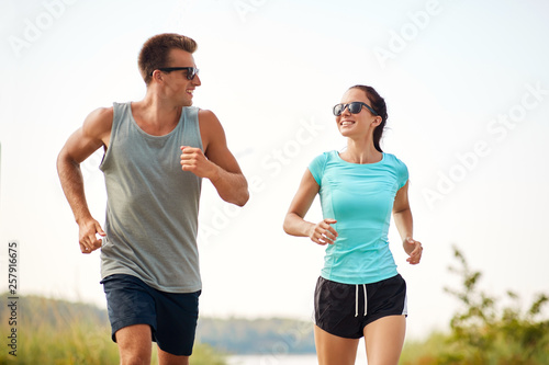 fitness, sport and lifestyle concept - happy couple in sports clothes and sunglasses running along summer beach