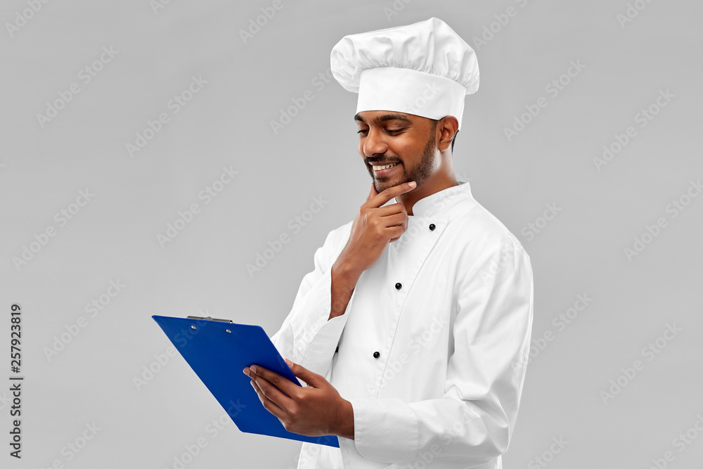 cooking, profession and people concept - happy male indian chef in toque reading menu on clipboard over grey background cooking, profession and people concept - happy male indian chef in toque reading menu on clipboard over grey background